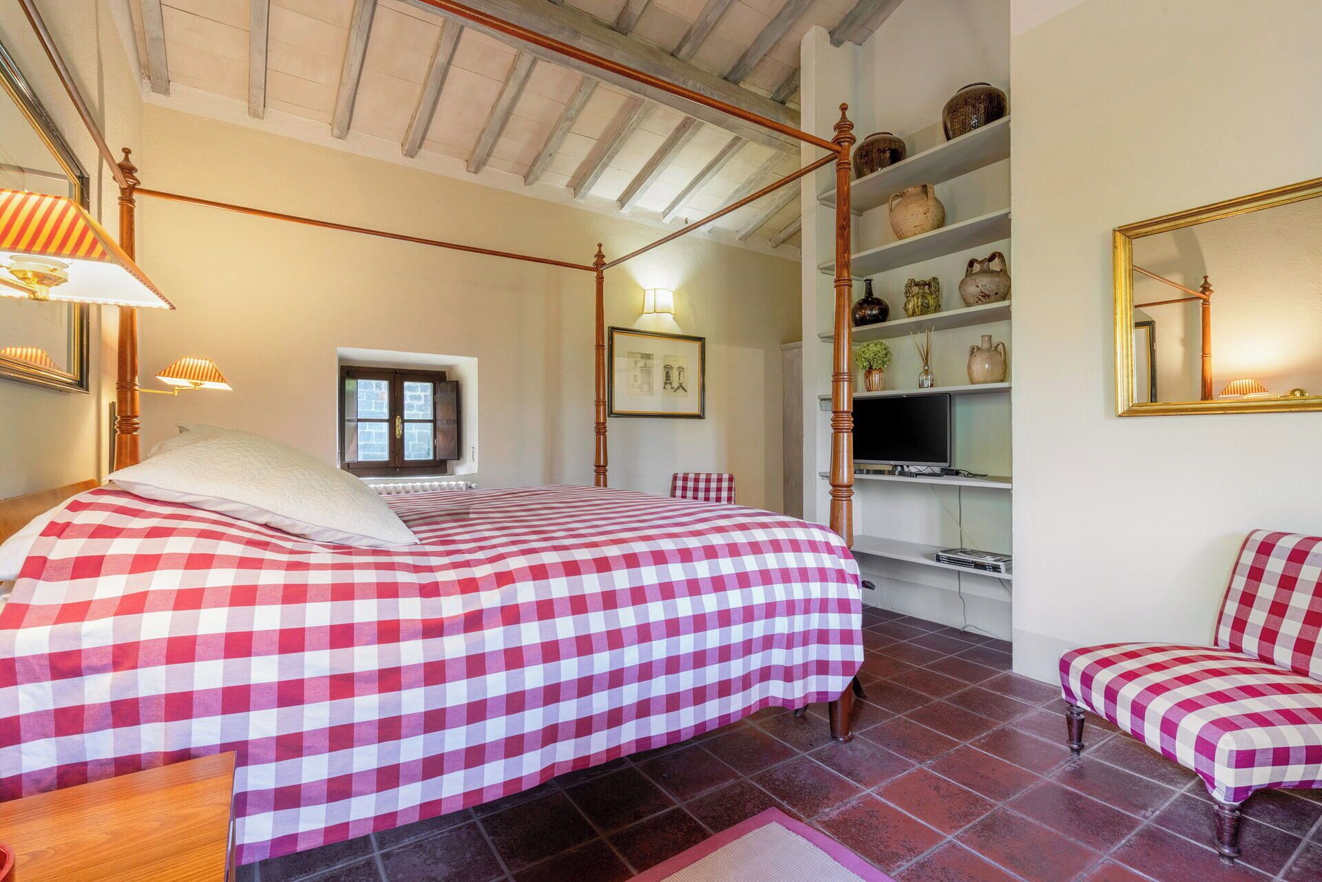Un dormitorio con cama con dosel, televisor, estantes con objetos decorativos y una ventana con contraventanas.