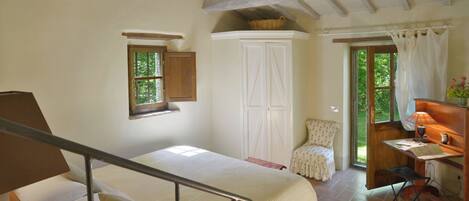 A bedroom with a bed, a small window, a wardrobe, a chair, a desk with a lamp, and a door leading outside.