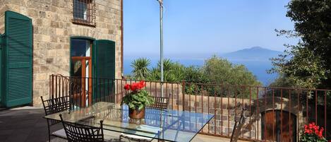 Eine Terrasse mit Glastisch und Stühlen, Blick auf das Meer und ein Steinhaus mit grünen Fensterläden.