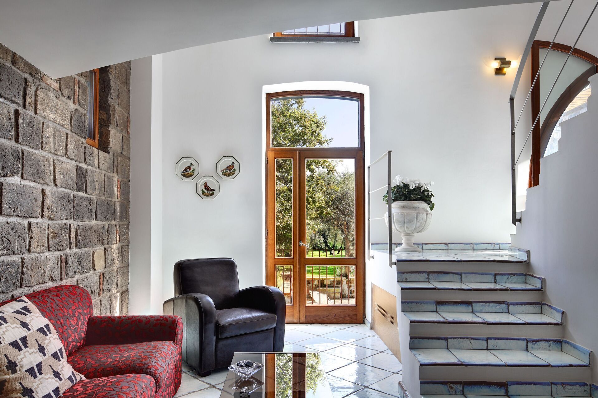 A spacious living area with a stone wall, a large wooden door leading to a garden, a staircase, and a seating area with a red patterned sofa and a black leather chair.