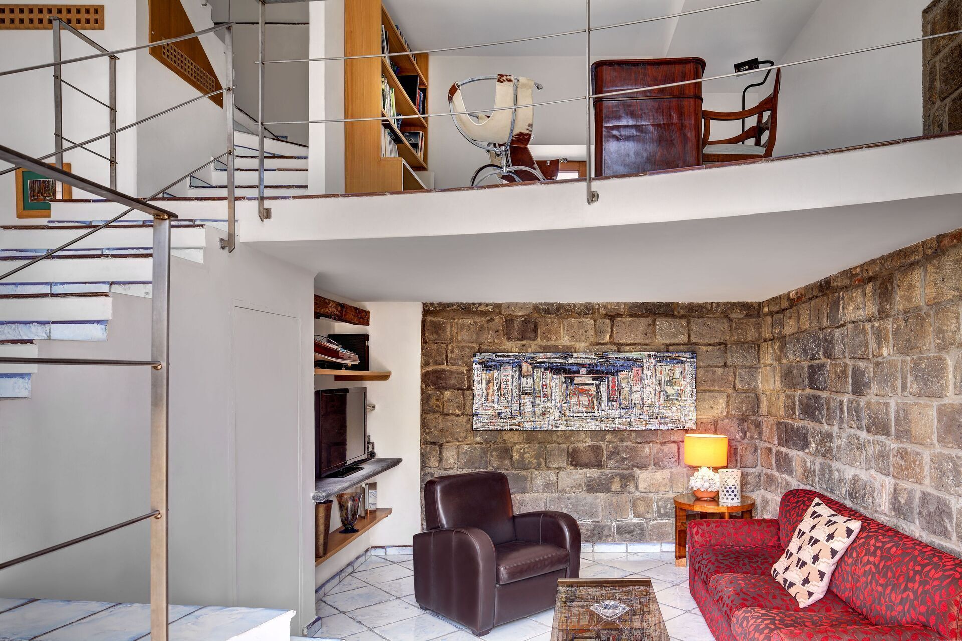 A spacious living area with a high ceiling, a stone wall, a red patterned sofa, a leather armchair, and a wooden bookshelf.
