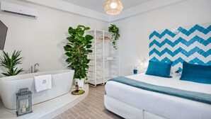 A modern bedroom with a white bed, blue pillows, a white wardrobe, a potted plant, and a bathroom with a bathtub and sink.