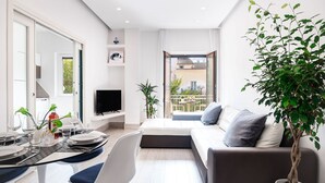 A modern living room with a sofa, a dining area with a table set for two, a flat-screen TV, and a balcony with a view of the city.