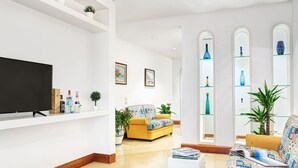 A modern living room with a flat-screen TV, a sofa, a yellow armchair, and a coffee table. There are potted plants and a bookshelf with decorative items.