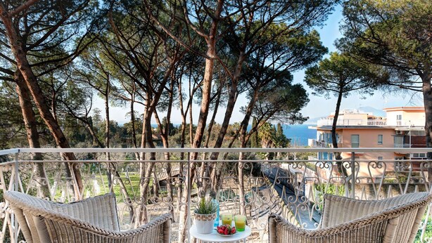 A balcony with wicker chairs, a small table with drinks and fruit, and a view of the sea and buildings.