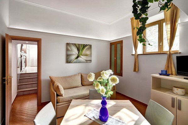 Una habitación con un sofá, una mesa de comedor con un florero, un televisor y una escalera que conduce a otra habitación.