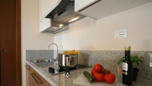 Una cocina con encimeras de granito, electrodomésticos de acero inoxidable, una olla de espagueti, tomates frescos, un pepino y una botella de aceite de oliva.