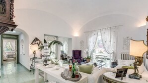 Un salón amplio con un sofá blanco, una mesa de centro y una araña. Se ve una ventana con cortinas traslúcidas y vistas al exterior.