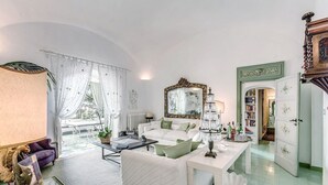 Un salón amplio con sofá, mesa de centro y una araña. Hay un espejo grande en la pared y una estantería llena de libros.