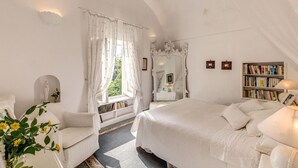 Un dormitorio con una cama grande, una mesita de noche, una silla, un ventanal con cortinas, un espejo, una estantería y una estatua.