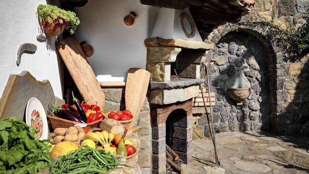 A rustic outdoor setting with a stone oven, a bell, and various vegetables and fruits displayed on a table.