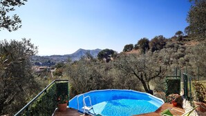 An outdoor swimming pool with a wooden deck, surrounded by olive trees and a view of a town and mountains in the background.