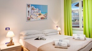 Habitación de hotel con cama, dos lámparas de noche, mesita de noche, ventana con cortinas verdes y un cuadro de una escena costera.