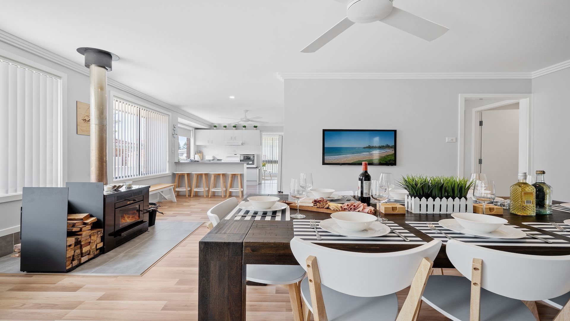 A modern kitchen with a central island, dining area with white chairs, and a large window with blinds.