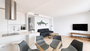 A modern kitchen with a stainless steel range hood, a dining area with a glass table and chairs, a dark grey sofa, and a flat-screen TV mounted on the wall.