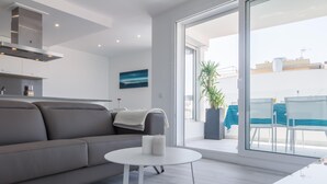 A modern living room with a grey sofa, a small white table, and a large sliding glass door leading to an outdoor area with a blue table and chairs. - 3-bedroom apartment with sea views, communal jacuzzi, next to the beach and close to the town center (Roses)