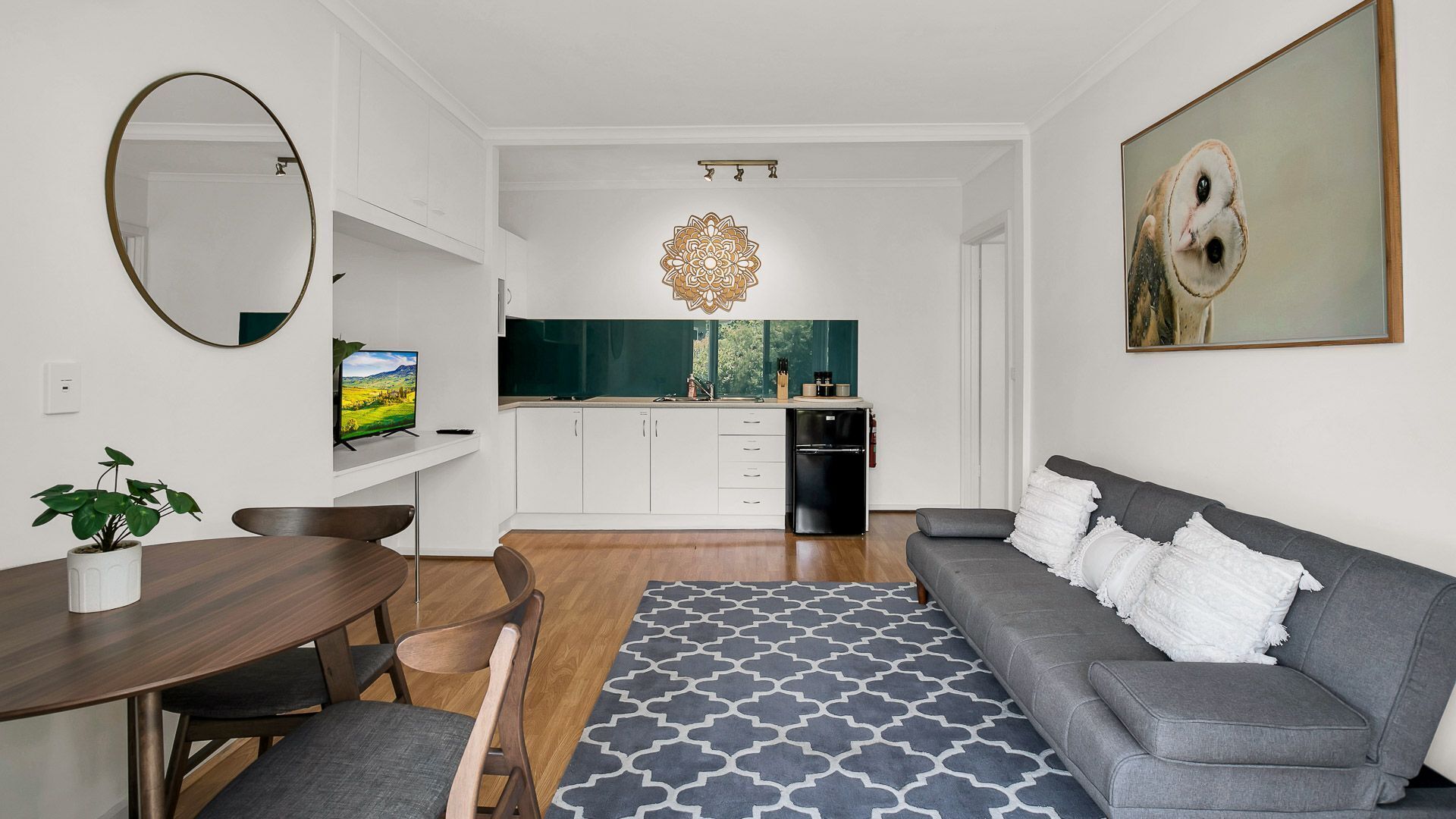 Ein modernes Wohnzimmer mit grauem Sofa, einem Esstisch mit Stühlen, einer Küche mit weißen Schränken und einem großen Spiegel an der Wand.