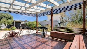 A covered outdoor patio with a barbecue grill, wooden deck, and a view of a modern building.