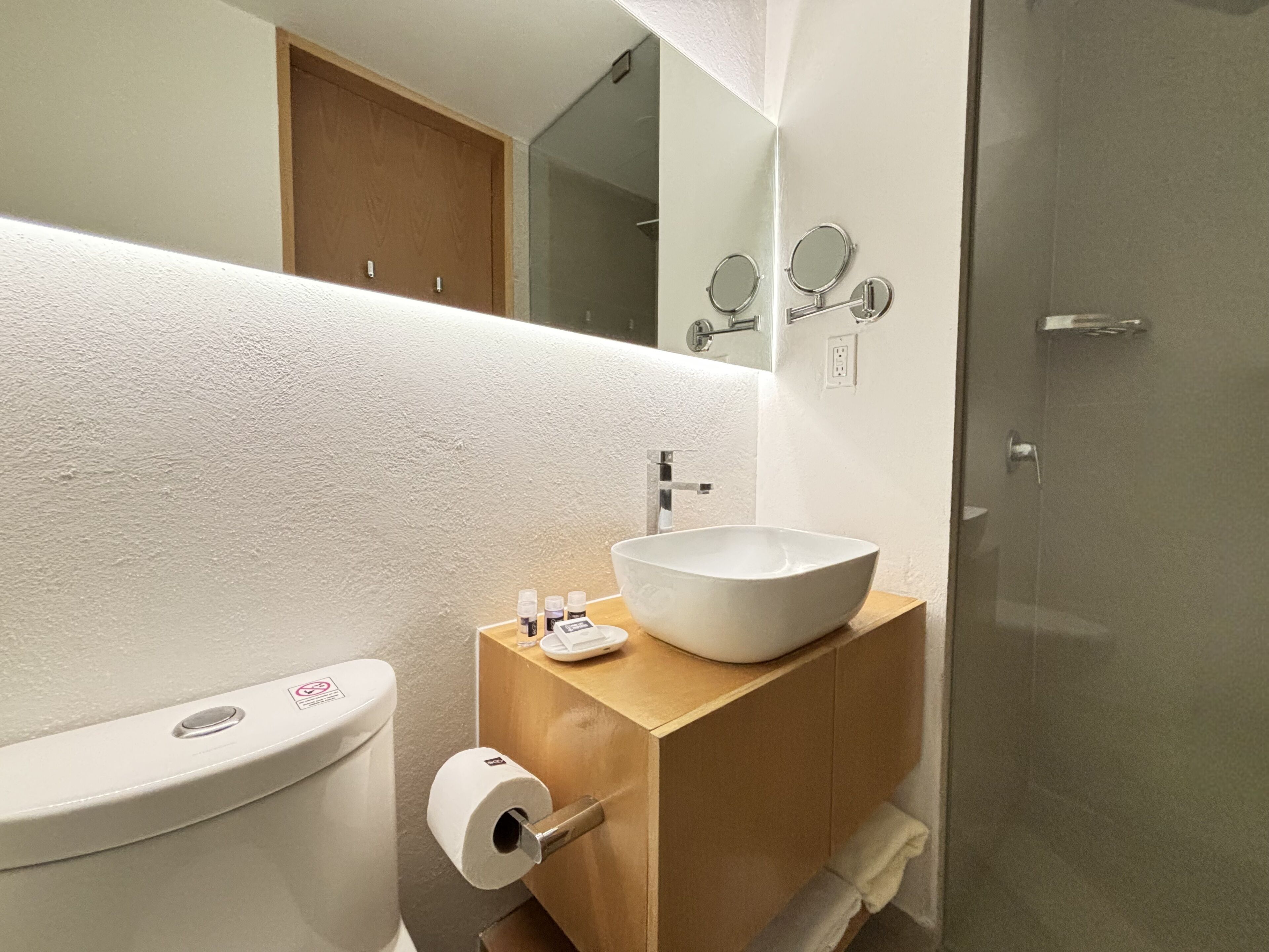 Un baño moderno con inodoro blanco, tocador de madera con lavabo blanco y cabina de ducha con mampara de vidrio.