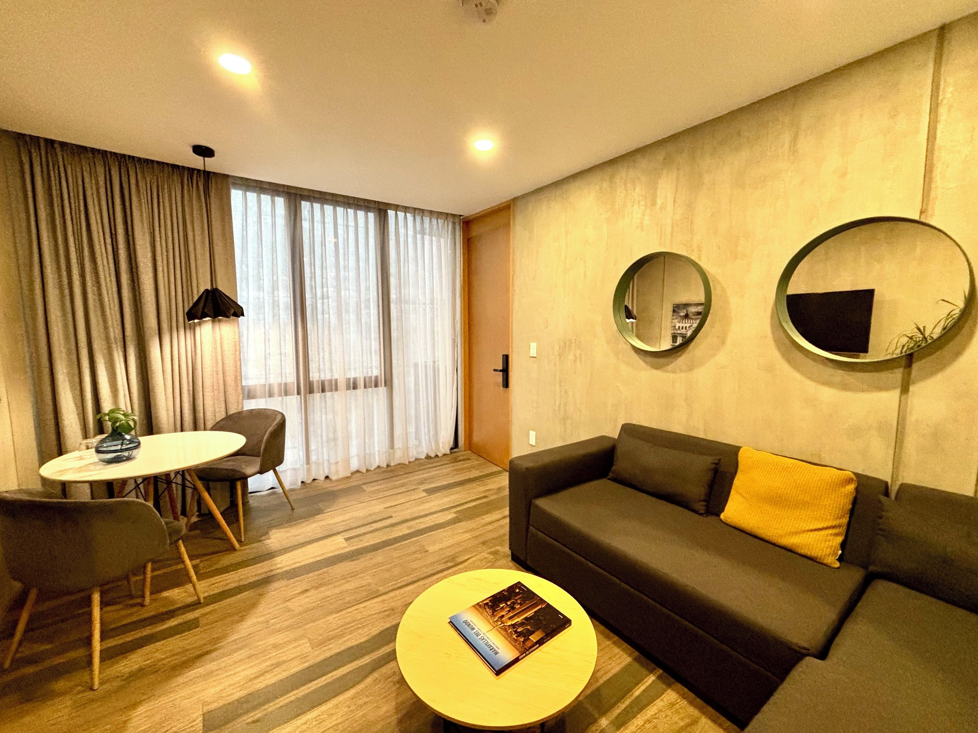 Ein modernes Wohnzimmer mit einer Couch, einem runden Couchtisch, einem Esstisch mit Stühlen und einem großen Fenster mit Vorhängen.