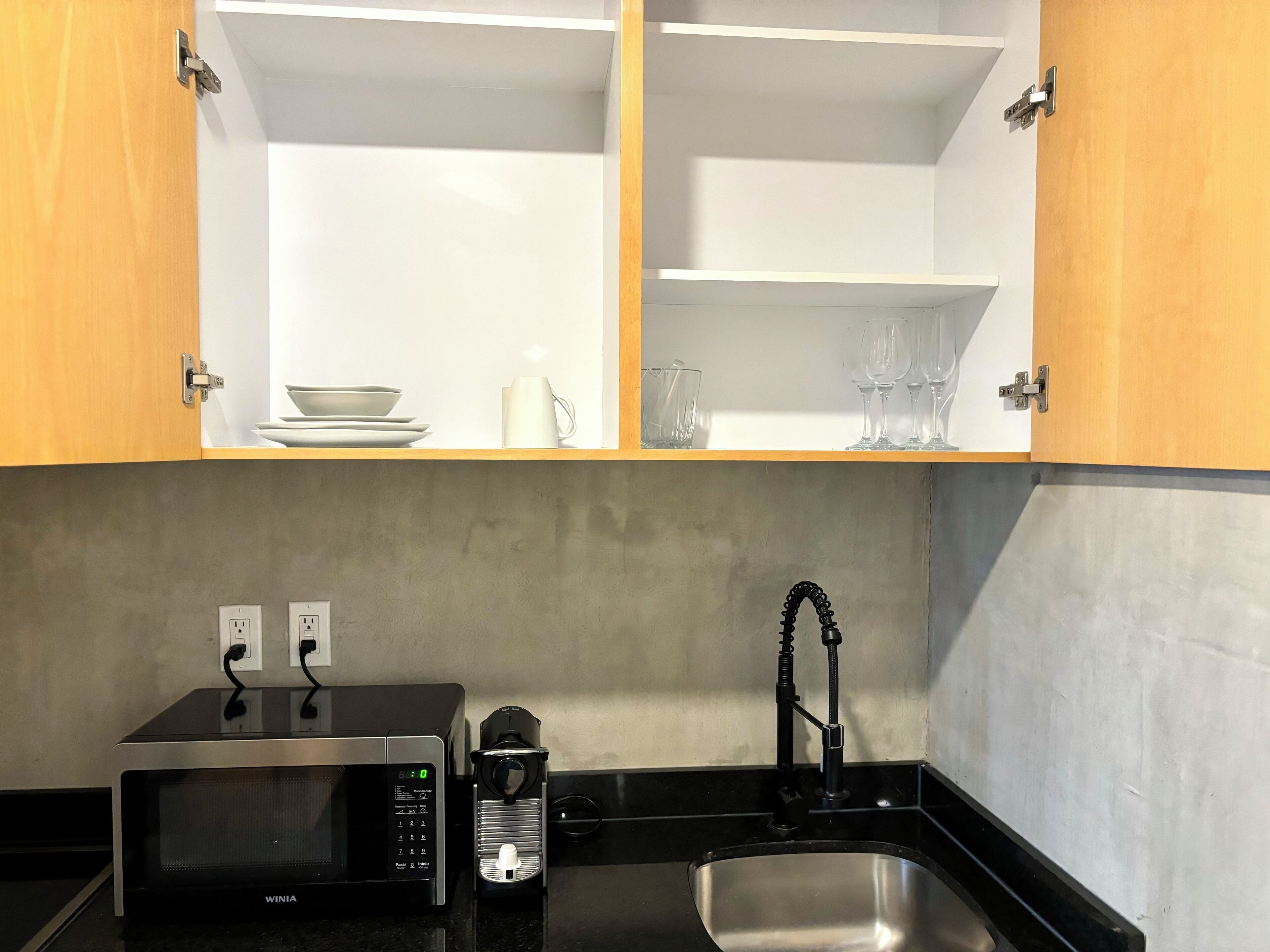 Una cocina equipada con microondas, cafetera, lavamanos y armarios de madera con estantes abiertos que contienen vajilla y vasos.