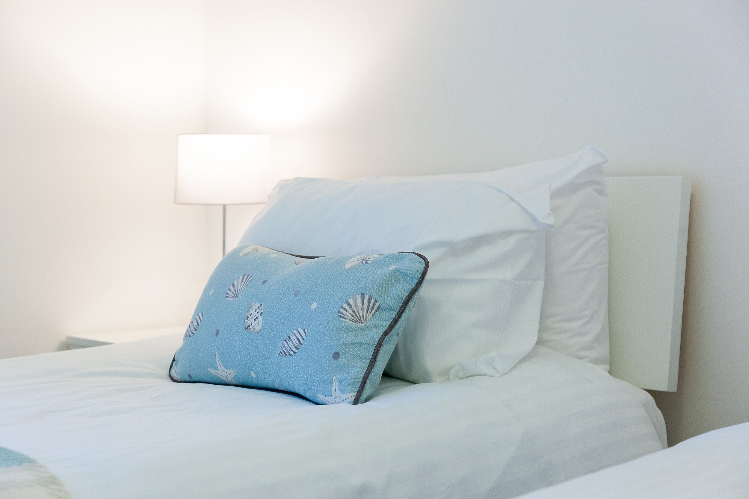 Una cama bien hecha con ropa blanca y una única almohada azul con estampado de concha.