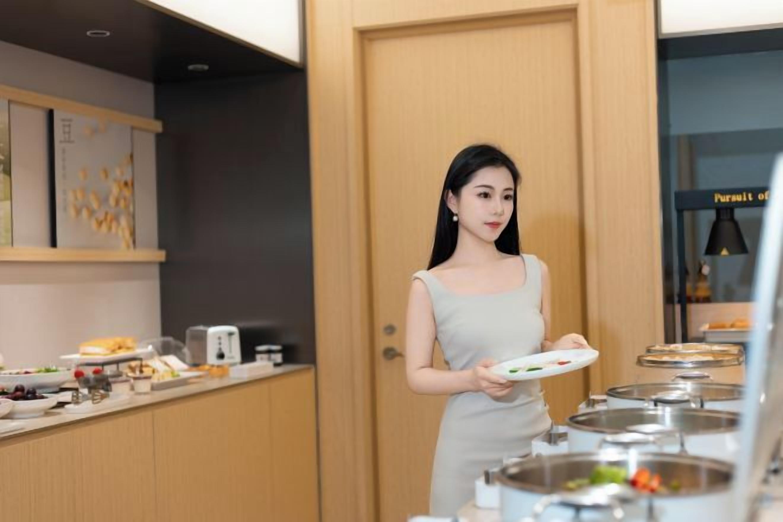 A woman in a light-colored dress holding a plate, standing in a room with a wooden door, a counter with food items, and a kitchen area with pots and a stove.