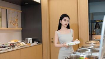 A woman in a light-colored dress holding a plate, standing in a room with a wooden door, a counter with food items, and a kitchen area with pots and a stove.