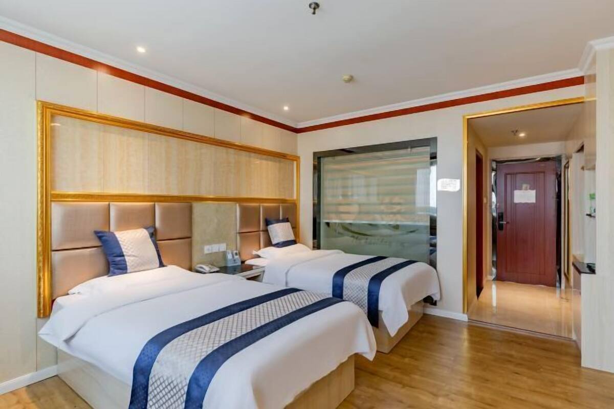 Habitació d'hotel amb dos llits, un panell de fusta al capçal, una separació de vidre i un paviment de fusta.