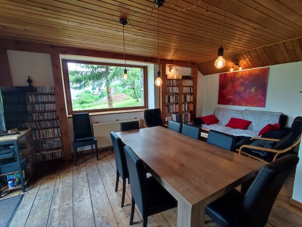 A cozy room with a wooden ceiling, a large window, a bookshelf, a dining table, and a sofa with red cushions.