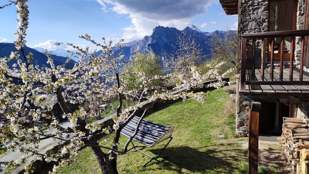 A stone building with a balcony overlooking a garden with blooming trees and a mountain backdrop.