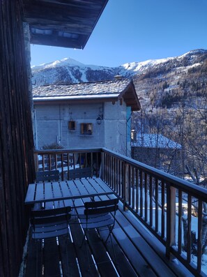 A wooden deck with two chairs, a view of snow-covered mountains, and a building with a tiled roof.