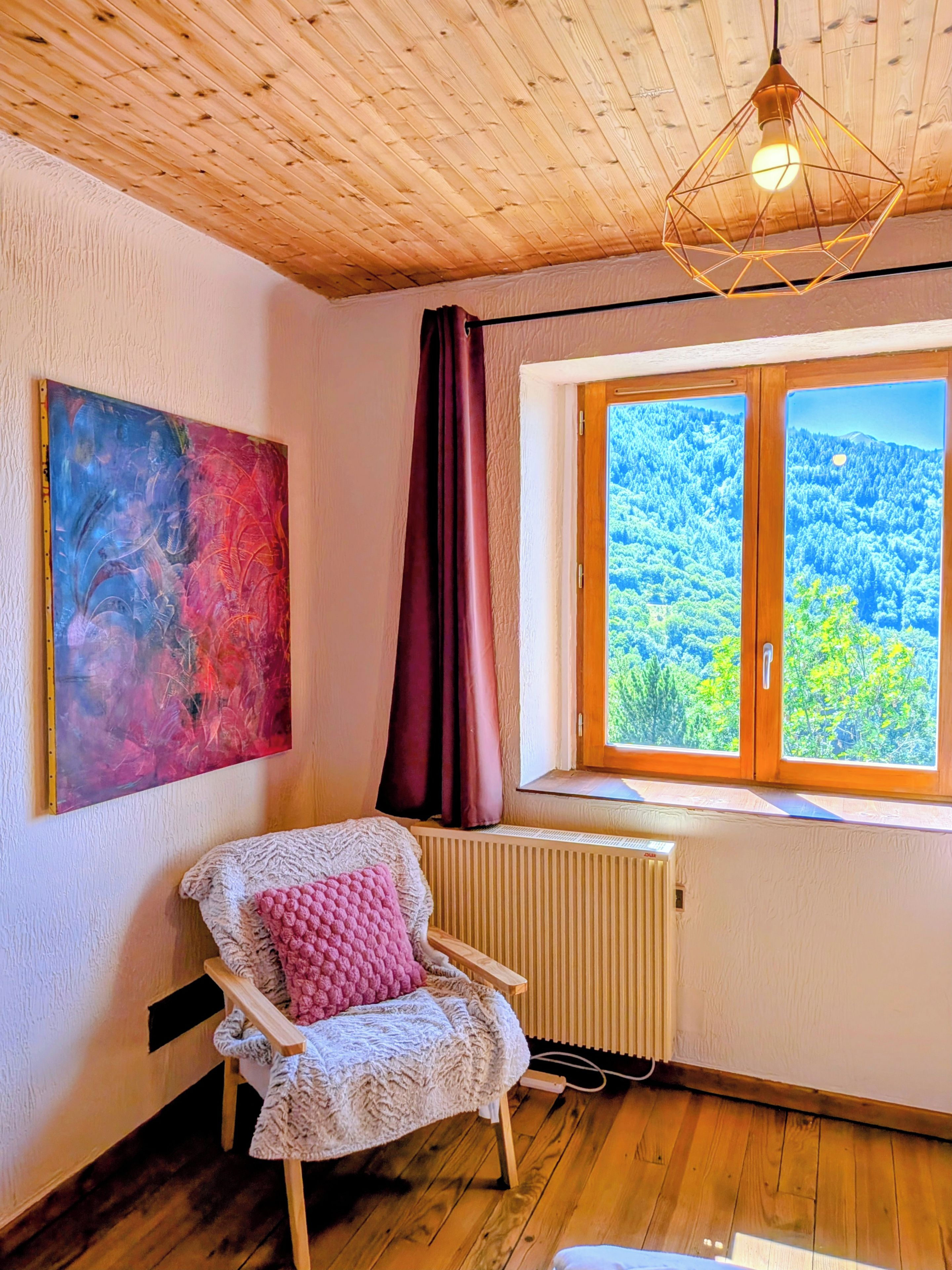 A room with a wooden ceiling, a radiator, a window with a view of a forest, a chair with a cushion, and a painting on the wall.