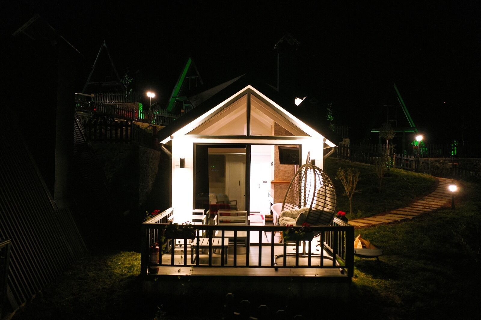 A small, well-lit house with a porch, a hanging chair, and a pathway leading to it.