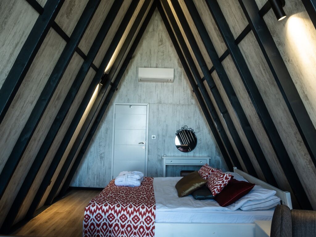 Une chambre avec un plafond en pente, un lit avec une literie à motifs, une table de chevet avec un miroir et un climatiseur fixé au mur.