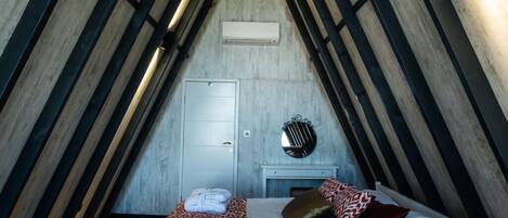 Une chambre avec un plafond en pente, un lit avec une literie à motifs, une table de chevet avec un miroir et un climatiseur fixé au mur.