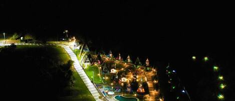 A nighttime aerial view of a resort with a winding road, multiple buildings, and a central pool area.
