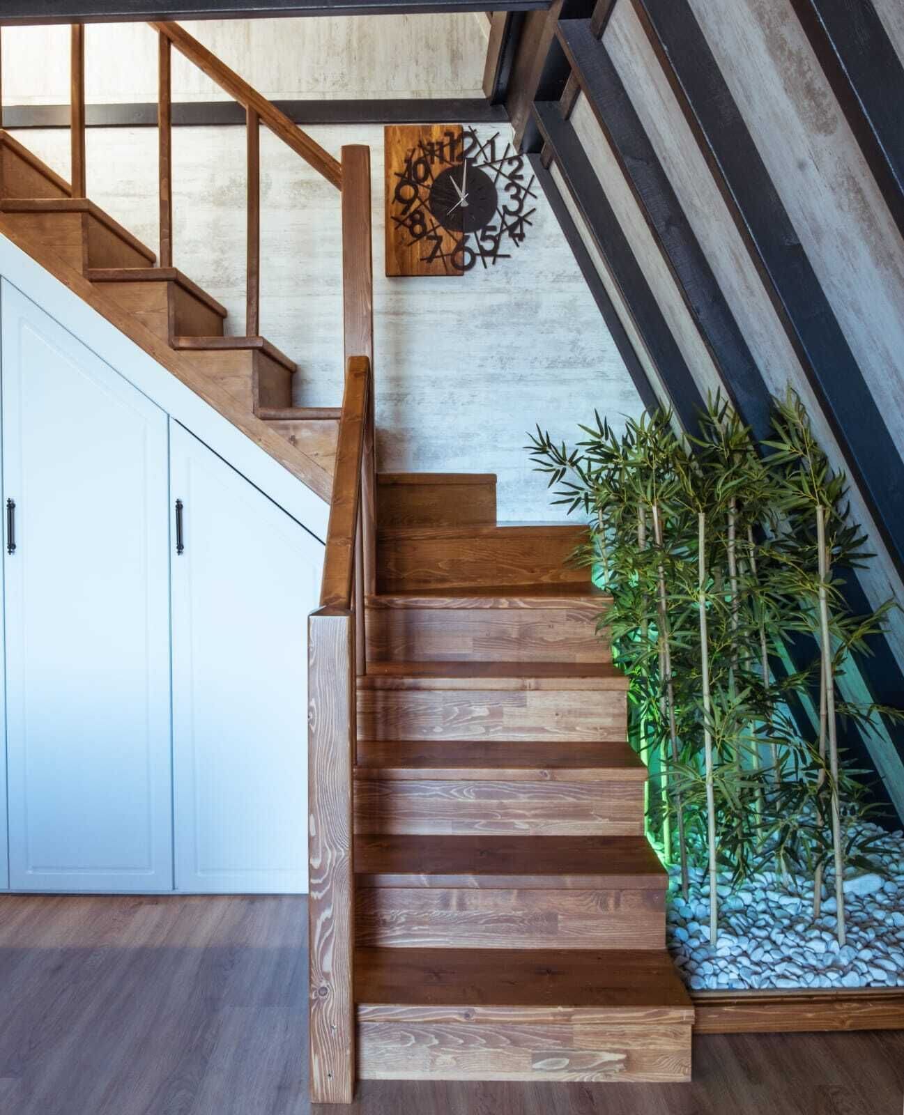 Un escalier en bois, avec une horloge au mur et une plante en bambou à côté.