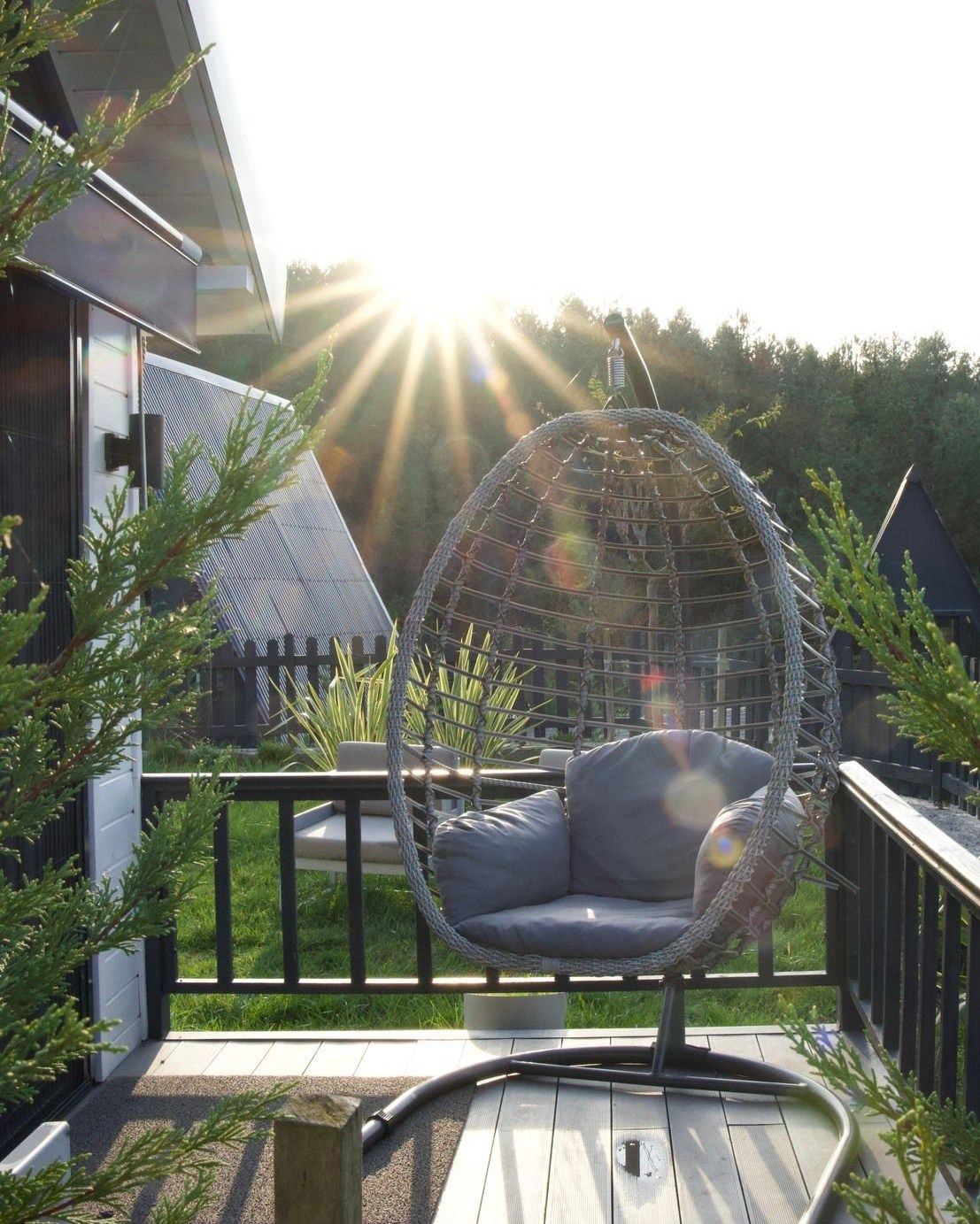 Una silla colgante en una terraza de madera con vista a una casa y árboles.