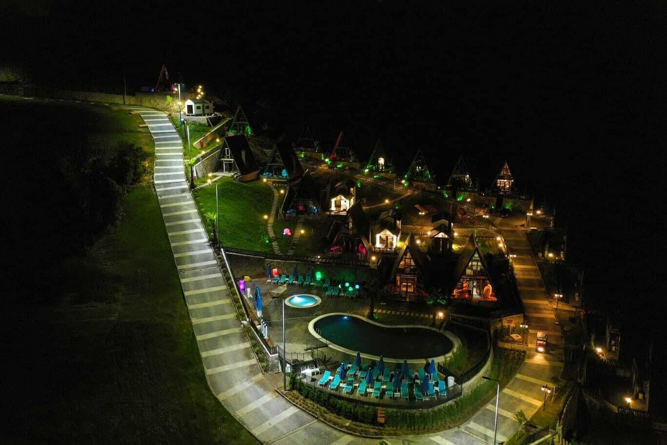 Luchtbeeld van een resort met een zwembad, ligbedden en verlichte gebouwen 's nachts.