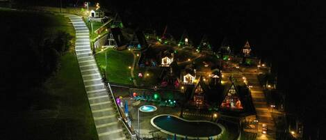 An aerial view of a resort with a swimming pool, lounge chairs, and illuminated buildings at night.