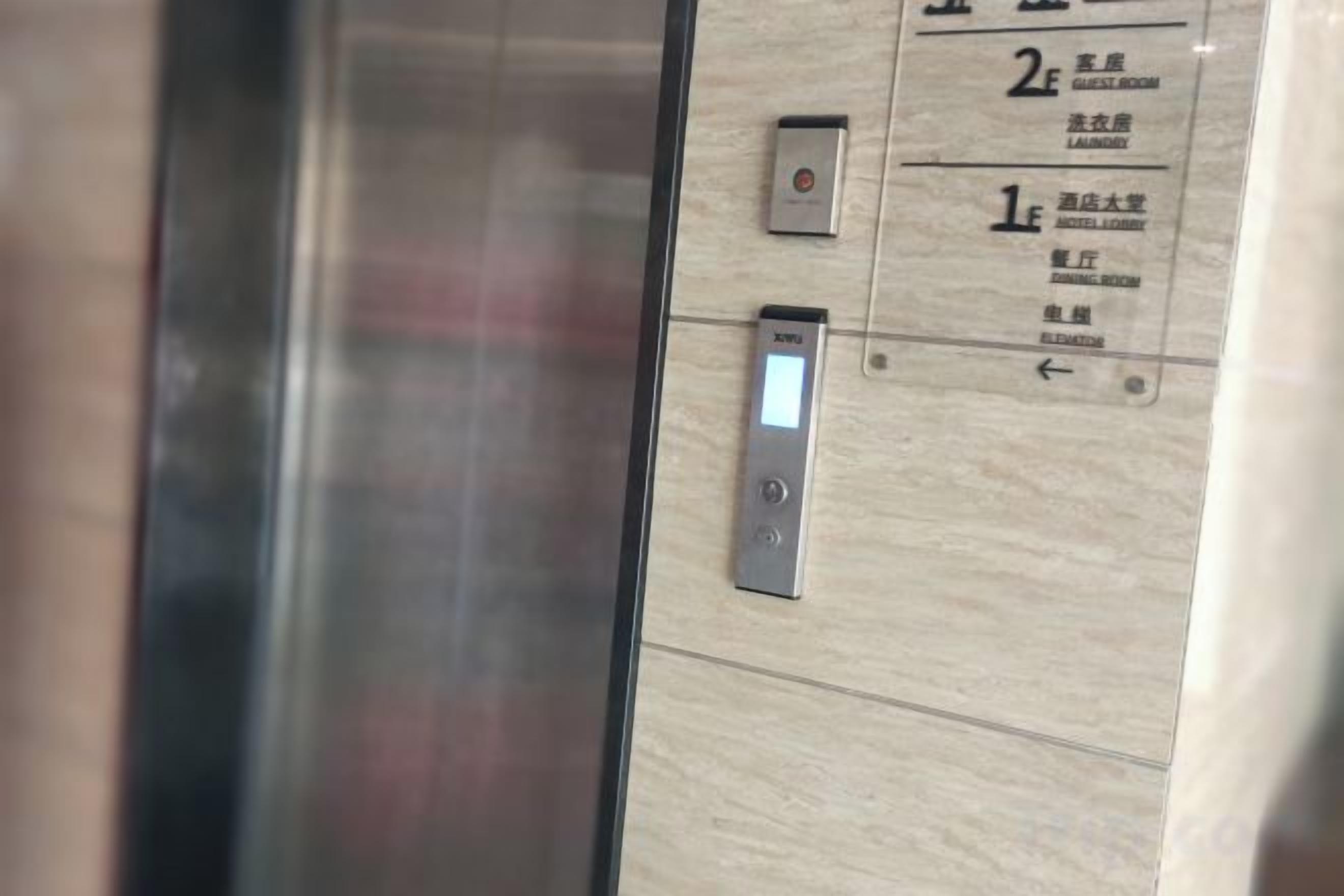 An elevator interior with a digital display and control panel, and a wall with room numbers and labels.