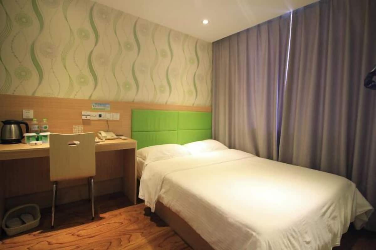 A hotel room with a green headboard, a bed with white linens, a desk with a chair, and a small table with a kettle and cups.