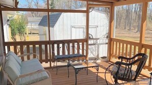 A covered outdoor area with wooden flooring, a ceiling fan, a cushioned chair, a metal table, and a view of a white building and trees. - Topridge - Spacious 3BR home w/fenced yard, pet friendly NO LINENS PROVIDED (Chincoteague)