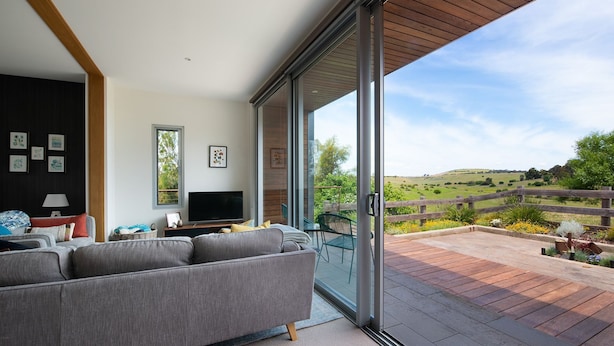 A modern living room with a large sofa, a flat-screen TV, and a view of a countryside landscape through a sliding glass door. - Kirkwood - Pet Friendly - Mid Century - Walking distance to Clunes! (Clunes)