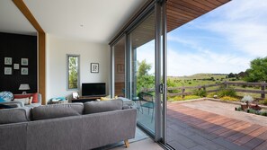 A modern living room with a large sofa, a flat-screen TV, and a view of a countryside landscape through a sliding glass door. - Kirkwood - Pet Friendly - Mid Century - Walking distance to Clunes! (Clunes)