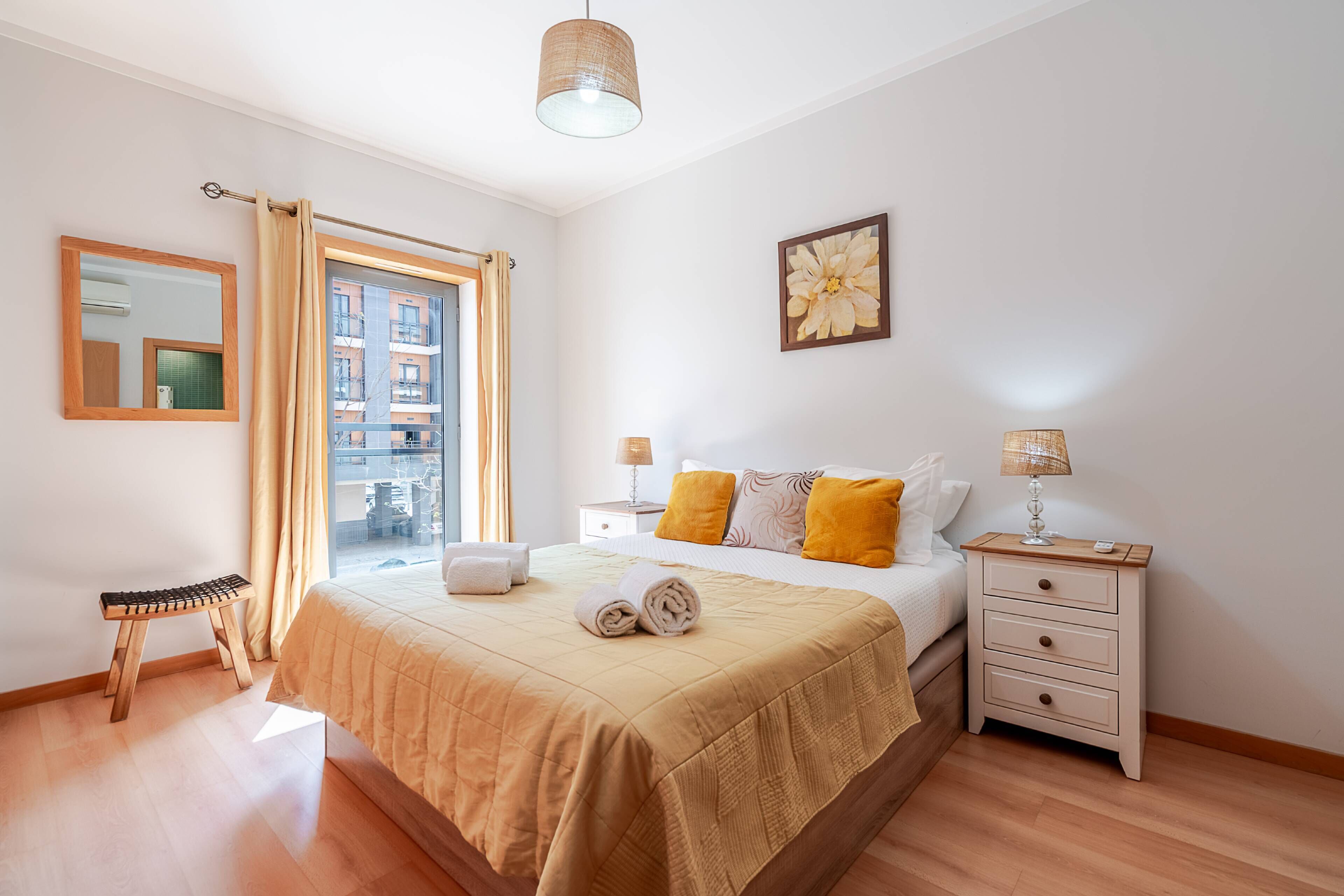 A hotel room with a large bed, a bedside table with lamps, a small stool, and a window with curtains.