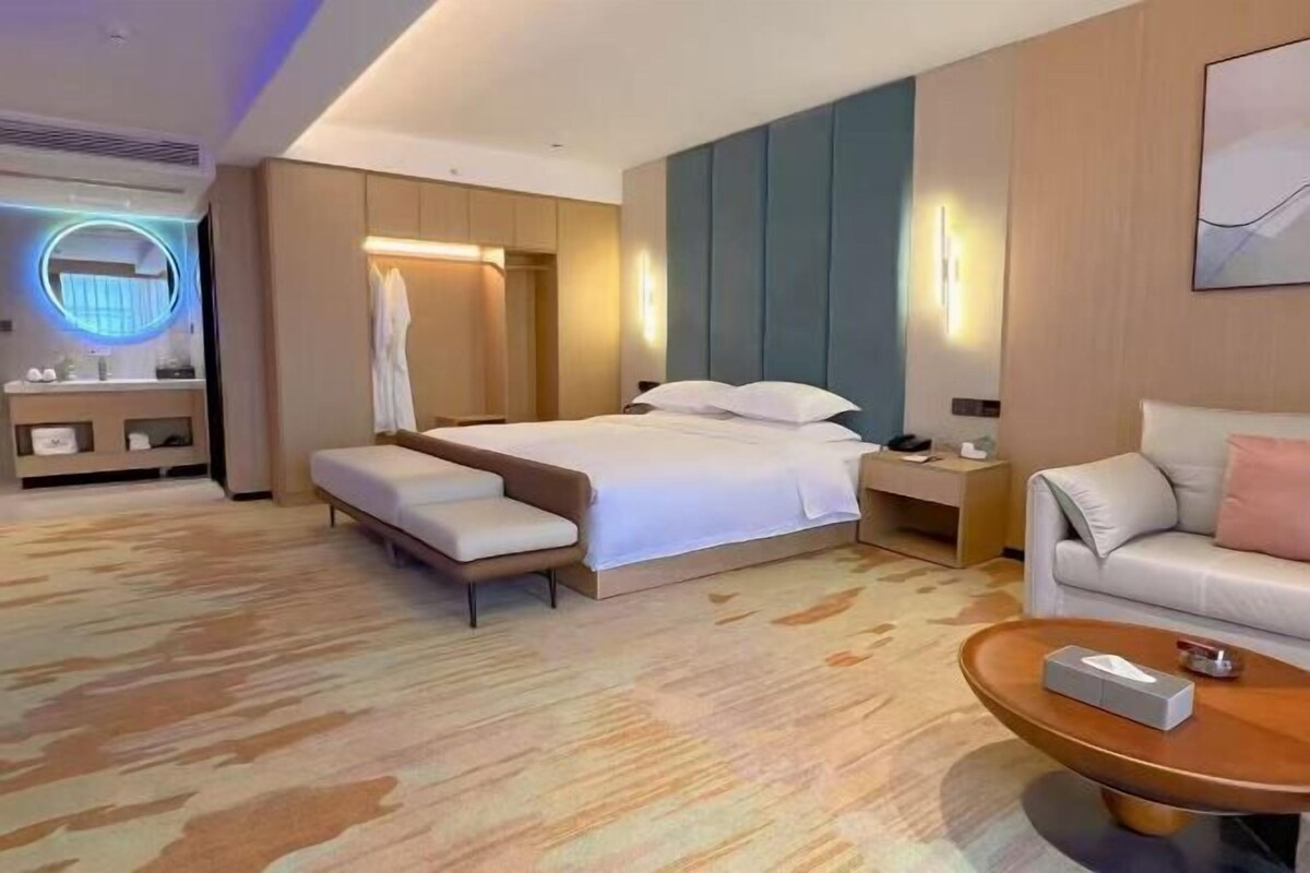 A modern hotel room with a large bed, a sofa, a round coffee table, and a bathroom with a mirror and towel rack.