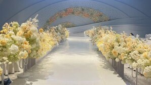Un couloir bordé de compositions florales, menant à une scène surmontée d’un luminaire circulaire bleu.