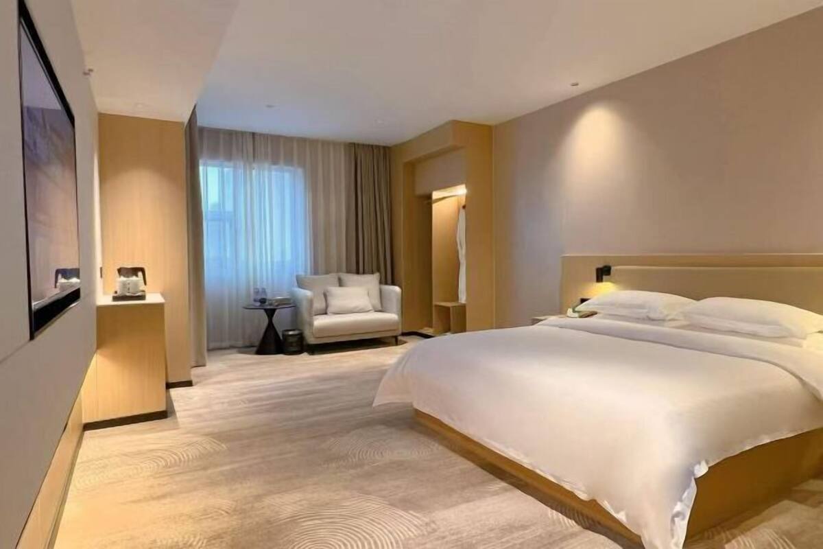 A hotel room with a large bed, a chair, a small table, and a window with curtains.